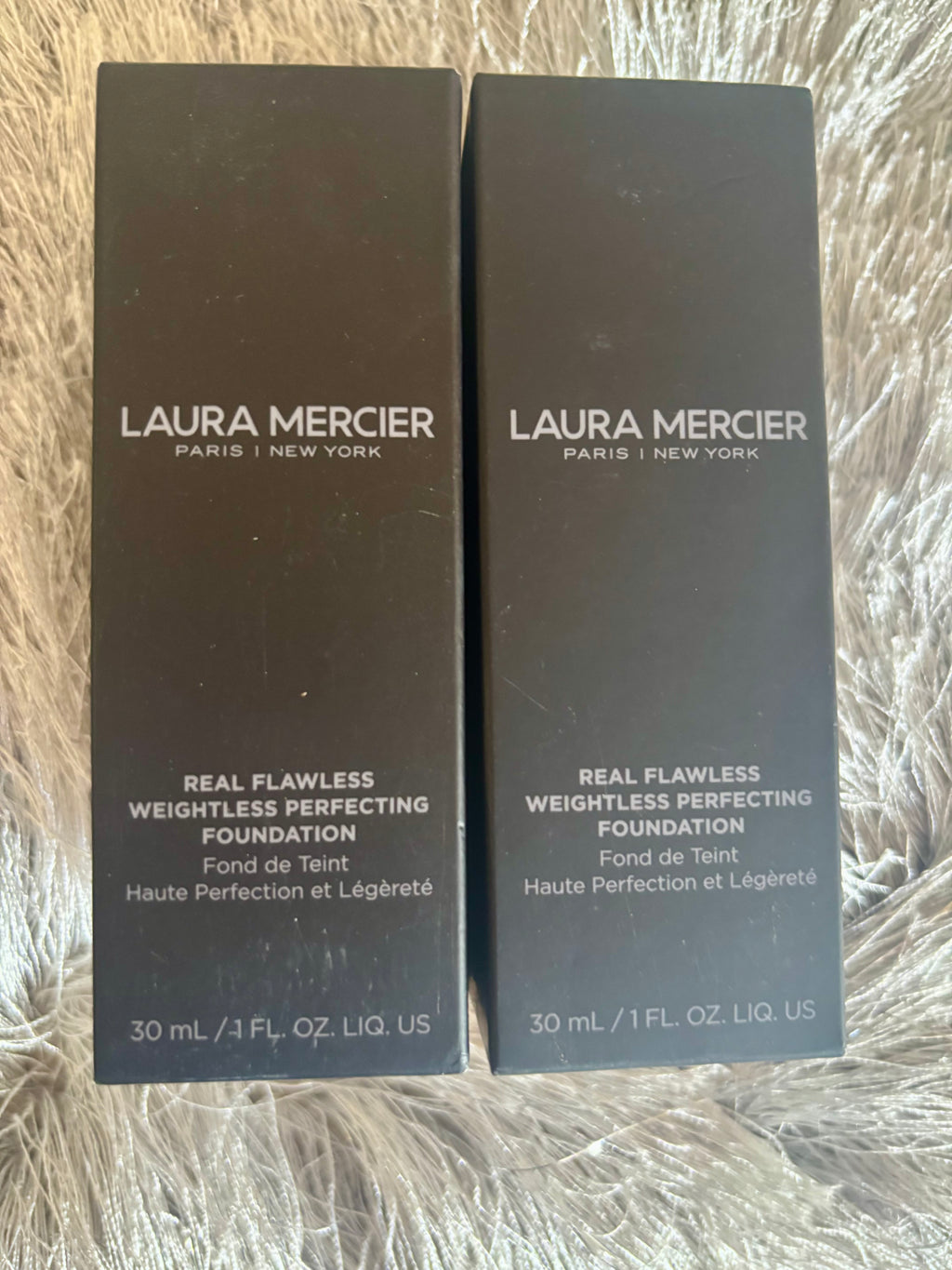 Two bottles of Laura Mercier Real Flawless Weightless Perfecting Foundation on a textured surface.